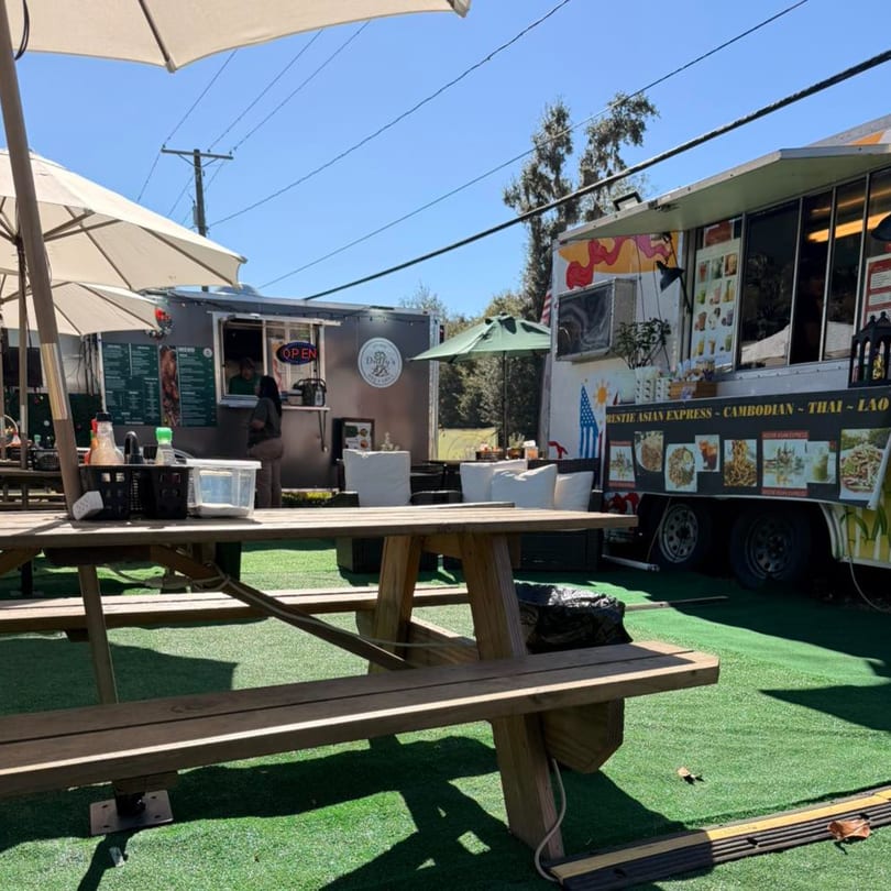 Outdoor seating area with picnic tables, umbrellas, and two food trucks under a clear sky.