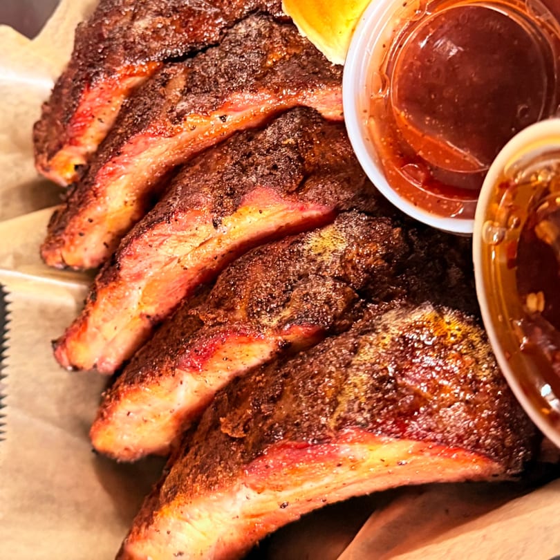 Close-up of Duffy's BBQ ribs with a smoky crust and two small cups of dipping sauce.