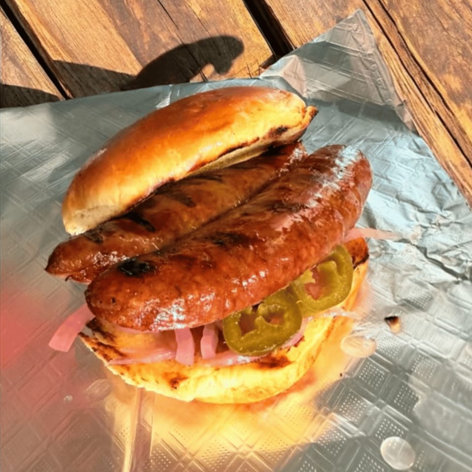 Two grilled sausages in a bun with pickled onions and jalapeños on foil, on a wooden table.
