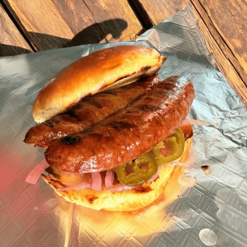 Two grilled sausages in a bun with pickled onions and jalapeños on foil, on a wooden table.