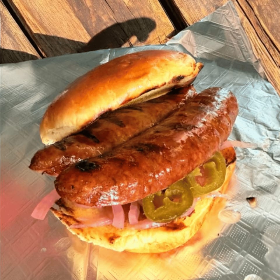 Two grilled sausages on a bun with pickled onions and jalapeños, on foil and a wooden table.