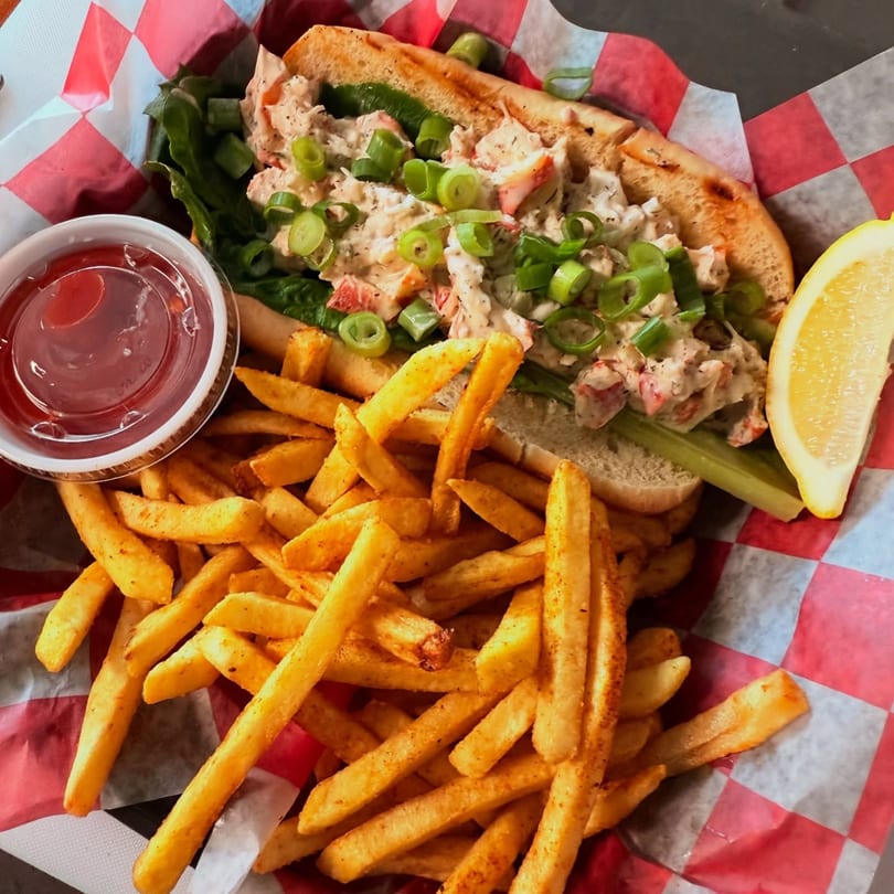 A lobster roll with fries, ketchup, and a lemon wedge in a red and white checkered basket.